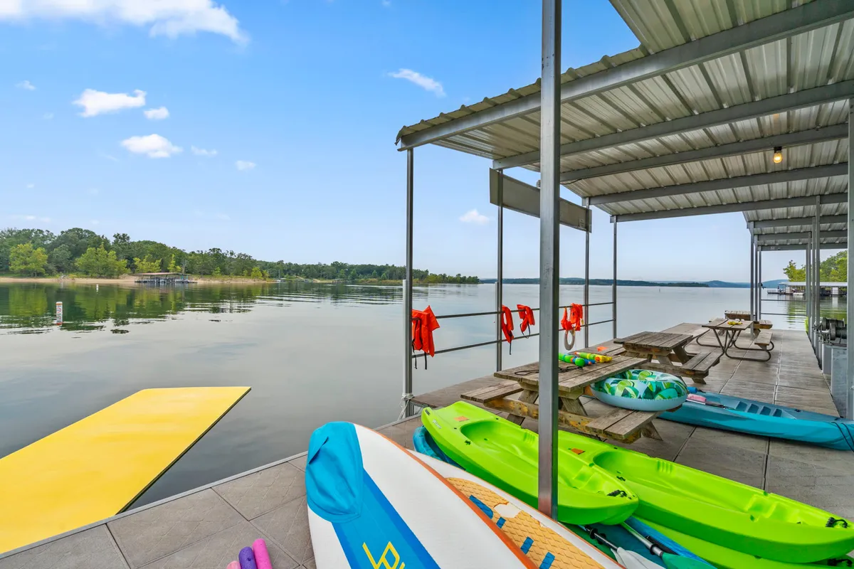 A colorful lakeside setup featuring kayaks ready for launch, a yellow floating mat, picnic tables, and life jackets by calm waters. The area looks inviting for sunbathing, swimming, kayaking, or simply relaxing by the lake.