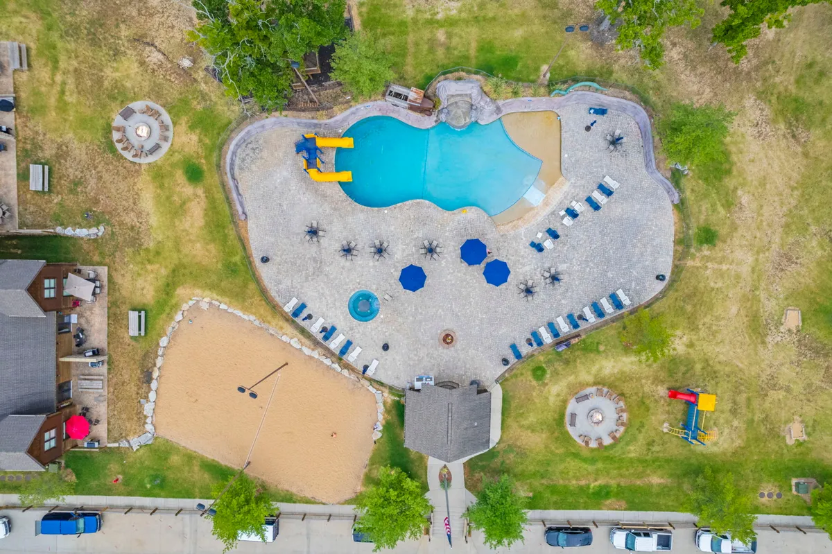 A vibrant outdoor pool area featuring a water slide, beach volleyball court, surrounding seating areas, and a fire pit. The sunny setting is filled with families enjoying swimming, sunbathing, and playing volleyball, while others gather around the fire pit.