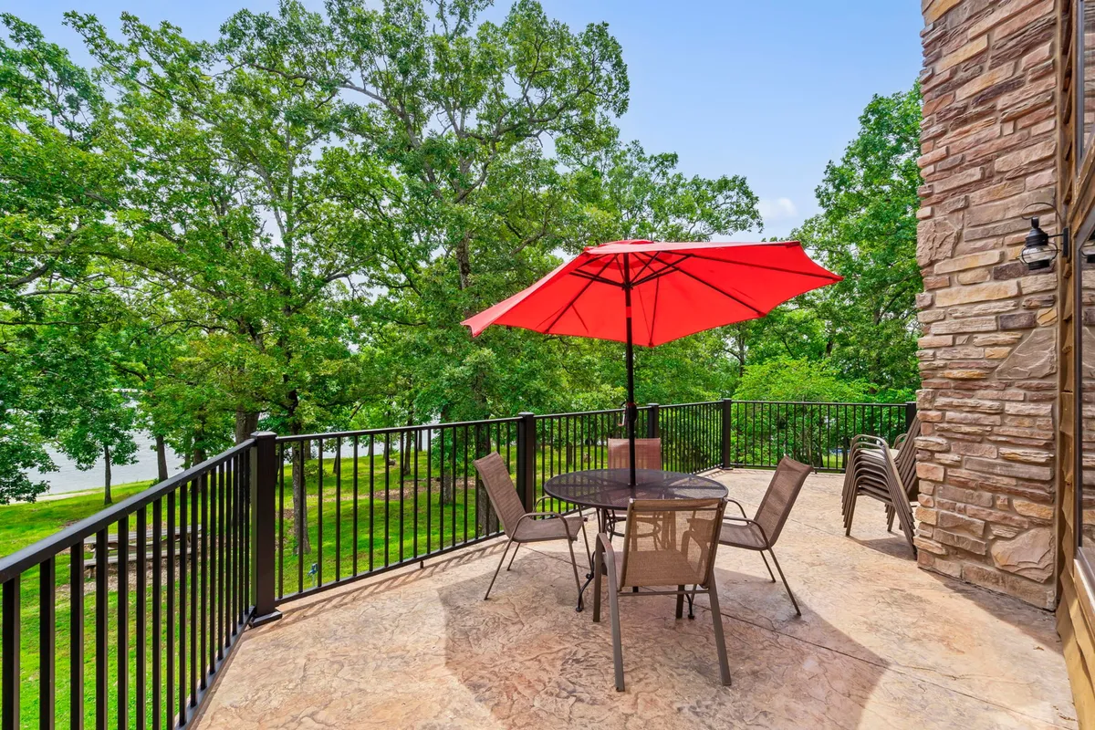This exterior shot features an outdoor dining set beneath a red umbrella, surrounded by lush greenery and overlooking the water. The scene is tranquil, inviting, and emphasizes outdoor leisure by the lake.