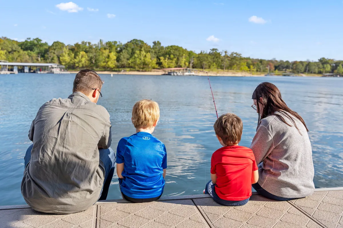 A family sits together at the edge of the calm lake, fishing rods in hand, surrounded by lush greenery. The mood is peaceful and joyful, highlighting a moment of family bonding as they anticipate catching fish and enjoy the natural setting.
