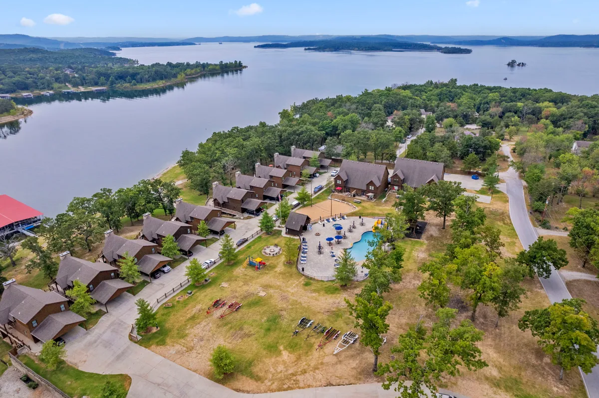An expansive exterior shot captures the lakeside swimming pool, surrounding cabins, lush green spaces, and the glimmering lake beyond. The image suggests a seamless connection between water activities, relaxation, and the natural setting.