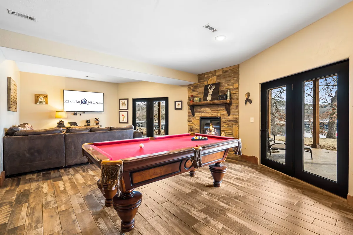 The image showcases a warm, inviting living area featuring a pool table as the centerpiece, a cozy fireplace, plush sofas for lounging, and expansive windows that flood the space with natural light and offer glimpses of the scenic outdoors. The setting conveys a perfect environment for guests to play pool, relax by the fire, socialize, and enjoy quality time together.
