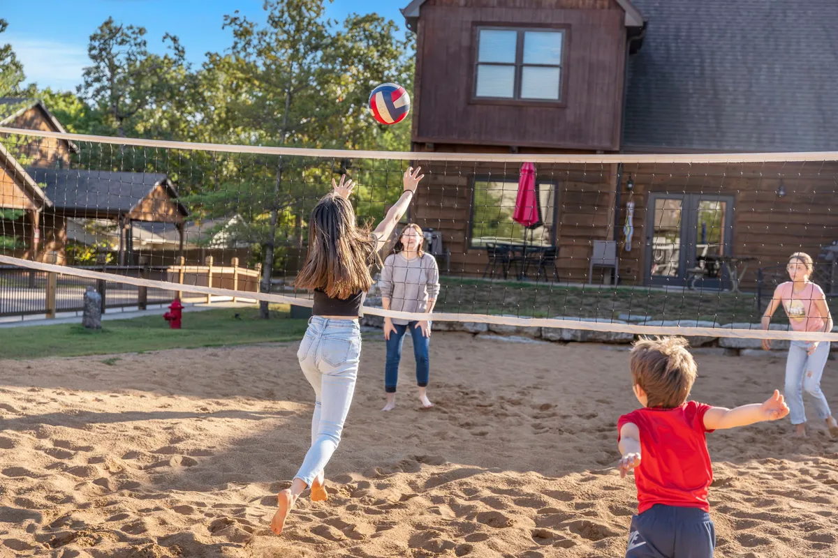 A joyful scene of a family volleyball game on a sandy court, with players actively engaged and a wooden house in the background. The setting sun casts a warm glow, adding to the vibrant, energetic mood of a family reunion or group gathering.
