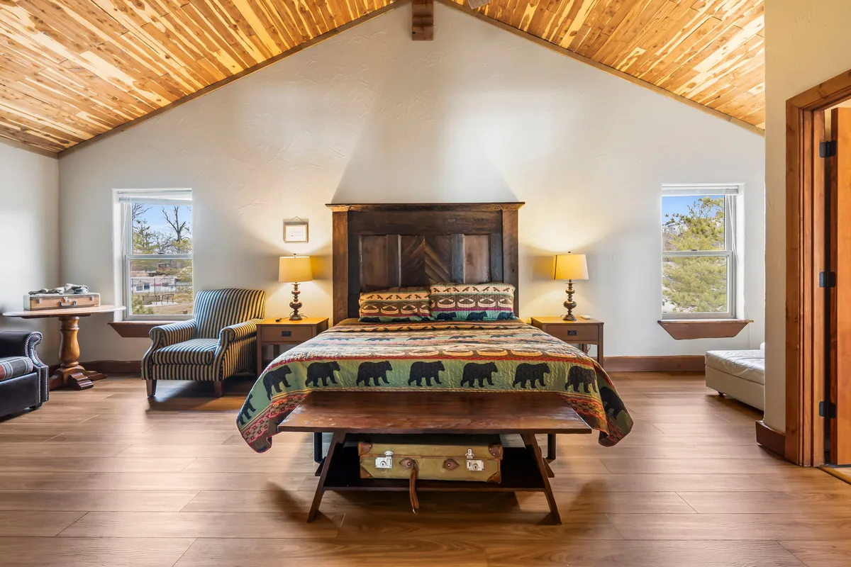 A warmly lit bedroom with a bear-patterned quilt on the bed, wooden headboard, two bedside lamps, and a striped armchair by the window. The room feels cozy and inviting, with natural light filtering in and comfortable furnishings designed for rest and relaxation.