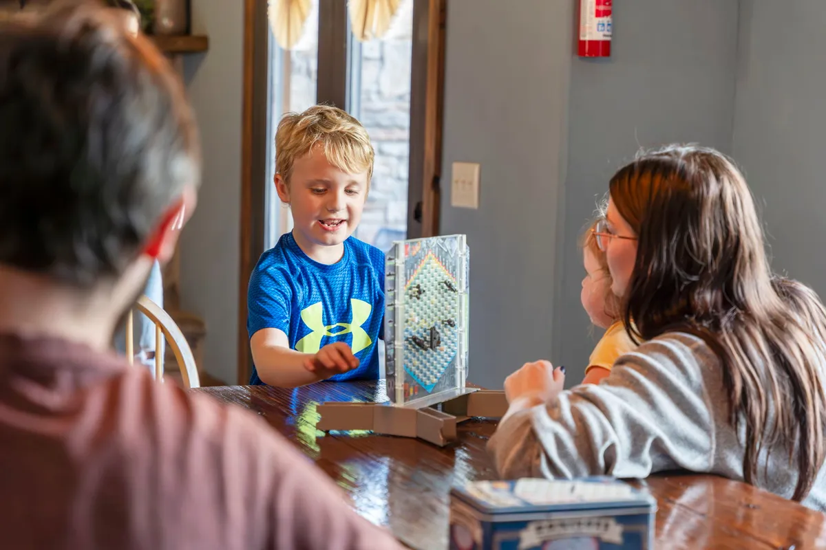 An interior scene of children engaged in a lively board game at a table, with a joyful, playful atmosphere. The scene highlights family bonding and friendly competition, reinforcing the idea of indoor entertainment during less favorable weather.