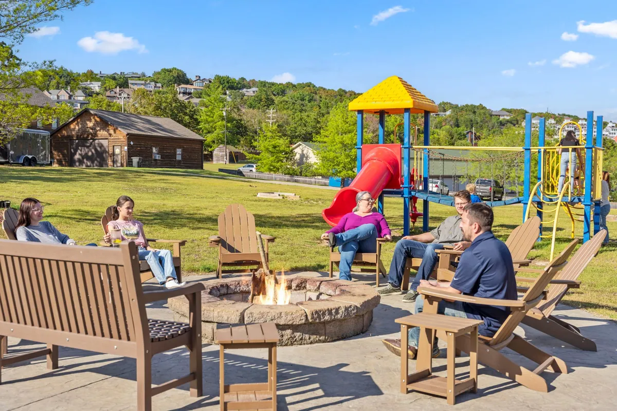 The image captures a warm, social setting around a fire pit, with people gathered and children playing on playground equipment in the background. The seating area and friendly atmosphere highlight the communal and relaxing aspects of the resort.