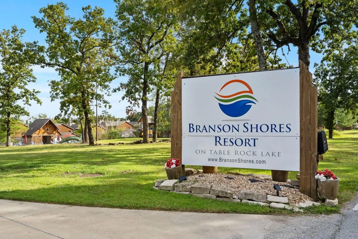 This image features the Branson Shores Resort sign surrounded by lush trees and nearby cabins, suggesting the welcoming arrival point for guests. The exterior scene hints at the family-friendly, lakefront atmosphere of the property and its proximity to both nature and Branson's attractions.