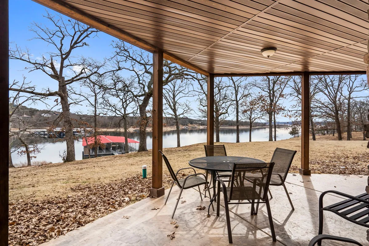 An exterior view of a peaceful lower-level patio with outdoor seating, lush greenery, and a beautiful lake view beyond. The covered patio offers a seamless transition between indoor play and outdoor relaxation.