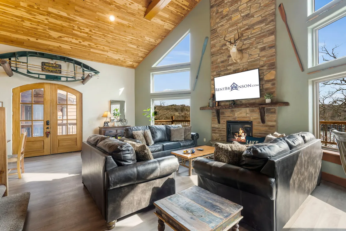 A spacious great room featuring a large stone fireplace with a TV above, plush leather sofas for relaxed seating, a wood-beamed ceiling, and a decorative canoe accent. Expansive windows offer sweeping lake views, flooding the room with natural light.