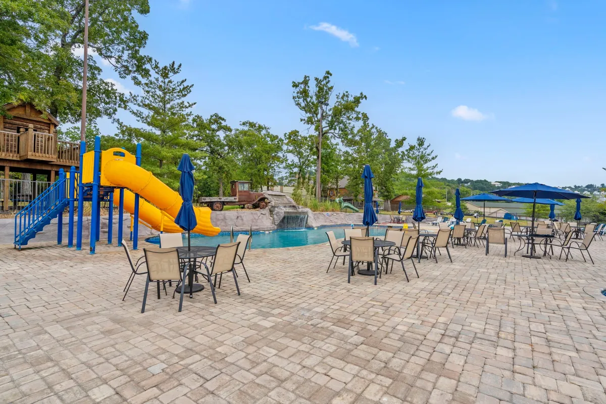 This image features a sparkling swimming pool with a water slide and a comfortable seating area, all set outdoors under sunny skies. The inviting water and nearby tables suggest both active fun and leisurely relaxation, capturing the essence of warm-weather enjoyment.