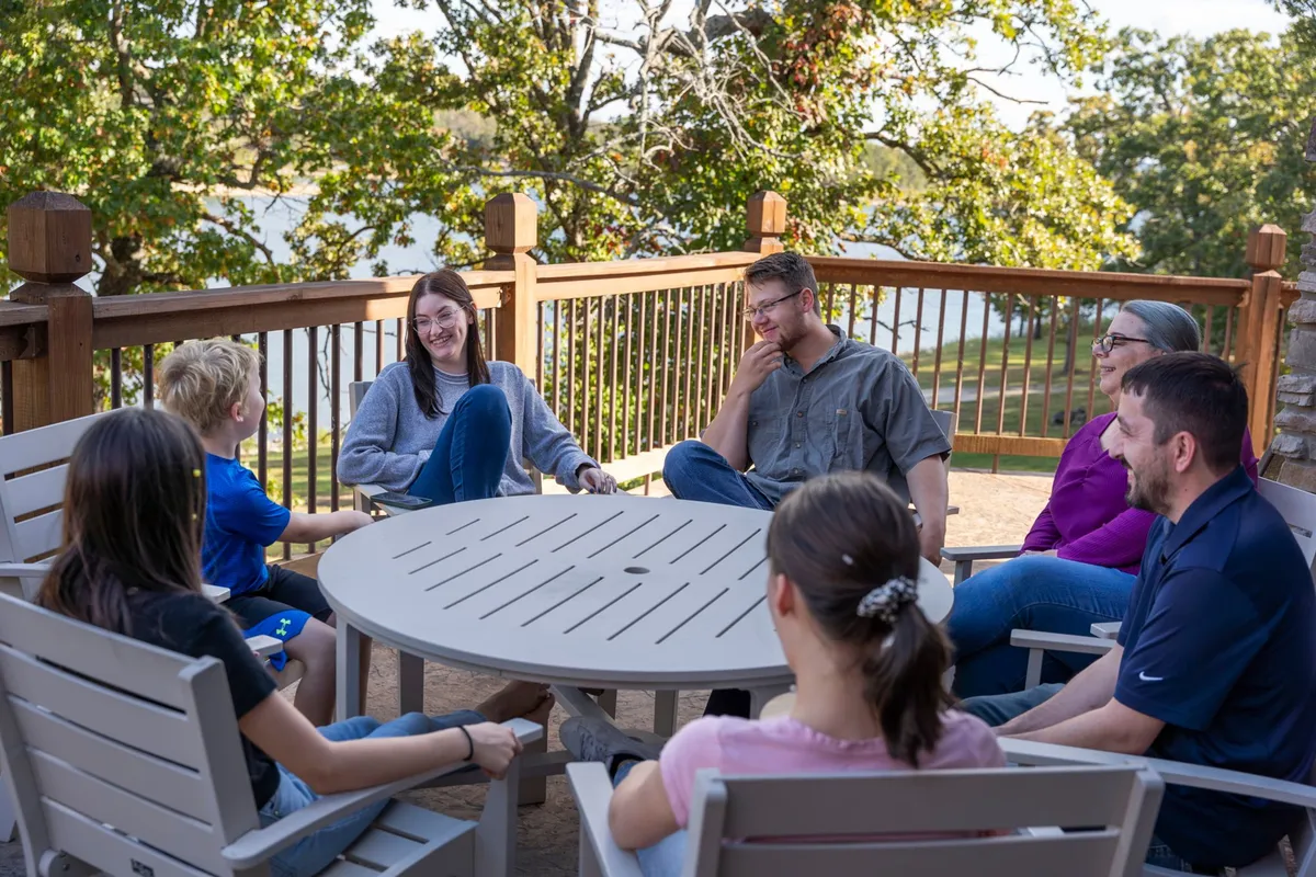 An exterior scene featuring a family gathered around a round table on a wooden deck, with a beautiful view of the lake in the background. The mood is relaxed and joyful, emphasizing togetherness, storytelling, and enjoying nature.