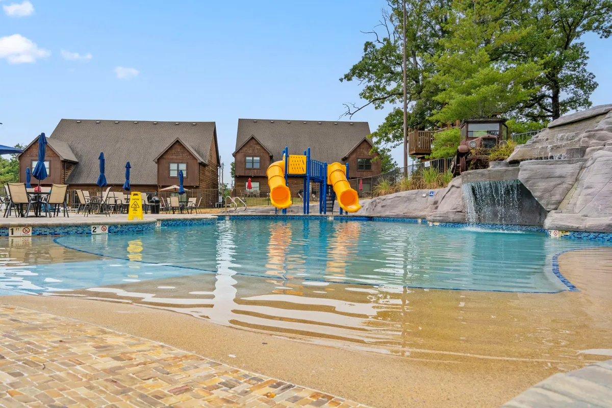 This vibrant exterior image features the resort’s swimming pool, complete with a water slide, waterfall feature, and surrounding cabins. Children and families are depicted enjoying the pool and poolside area, conveying a playful, social, and relaxing atmosphere.