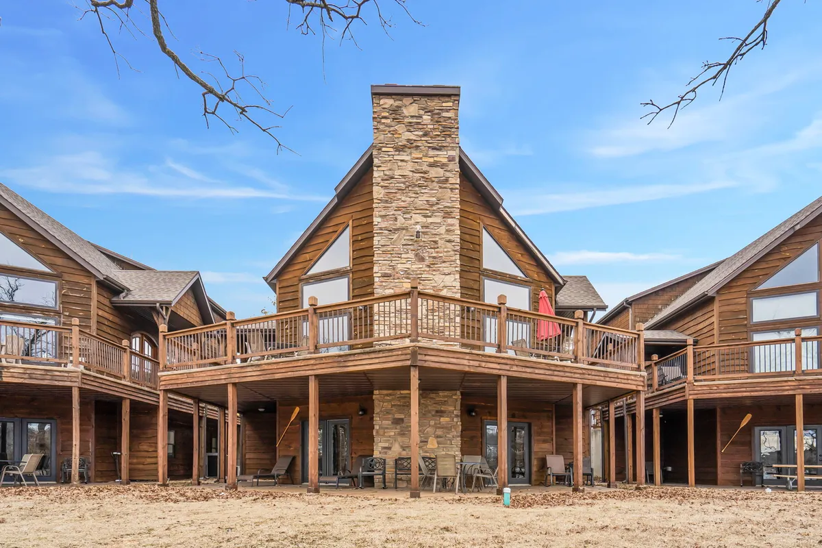This exterior image showcases a welcoming wooden deck with stone chimney, large windows, and comfortable outdoor furniture, all overlooking a scenic sunset. The setup is clearly designed for group gatherings, barbecues, and relaxation, emphasizing the spaciousness and beauty of the outdoor entertaining spaces.