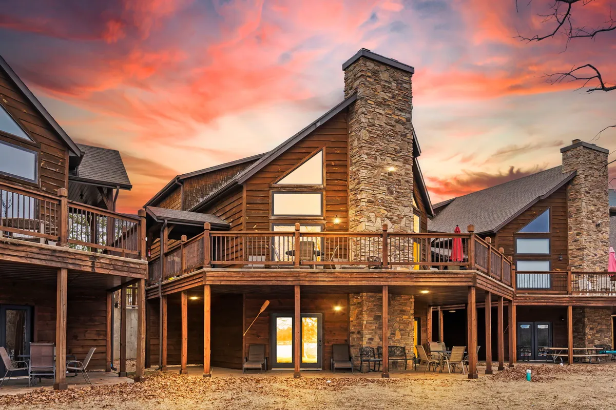 This image captures the exterior of the resort, featuring a spacious wooden deck with a stone chimney, large windows, and a warm sunset sky. The outdoor setting is welcoming and spacious, ideal for relaxing with a pet and enjoying the surrounding nature.